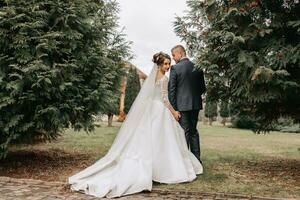 Stylish bride and groom tenderly embrace and kiss. Beautiful bride with modern bouquet embracing stylish groom in autumn park. Sensual romantic moment. Wedding. Stylish and beautiful. photo