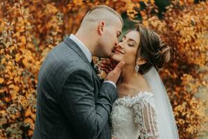 Stylish bride and groom tenderly embrace and kiss. Beautiful bride with modern bouquet embracing stylish groom in autumn park. Sensual romantic moment. Wedding. Stylish and beautiful. photo