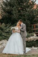 Stylish bride and groom tenderly embrace and kiss. Beautiful bride with modern bouquet embracing stylish groom in autumn park. Sensual romantic moment. Wedding. Stylish and beautiful. photo