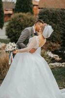 Stylish bride and groom tenderly embrace and kiss. Beautiful bride with modern bouquet embracing stylish groom in autumn park. Sensual romantic moment. Wedding. Stylish and beautiful. photo