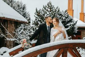 happy bride and groom embrace among snowy trees on a wooden bridge. Groom and bride in the winter park. Bride with a bouquet of flowers in a wedding dress and poncho. Groom in a black coat. photo