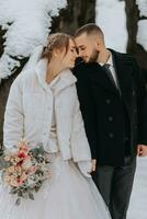 Wedding in winter. A stylish bride in a white mink fur coat with a bouquet of flowers in her hands and a groom in a black coat in a forest with white snow in cold weather. photo