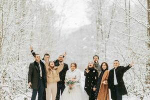 Happy newlyweds and their friends in winter coats stand in a snow-covered forest and throw snow with their hands photo