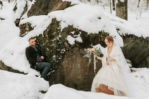 Wedding in winter. Stylish bride in white mink coat and groom in black coat in forest with white snow in cold weather. The bride and groom are walking among rocks and large stones in the forest photo