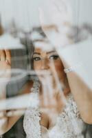 un hermosa y amable novia mira fuera el ventana en el Mañana. foto mediante el ventana. hermosa natural constituir. Boda peinado. Mañana de el novia.