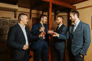el novio y su amigos en elegante trajes bebida whisky en el hotel habitación, el Mañana antes de el Boda preparativos. grupo de hombres hablando y Bebiendo whisky foto
