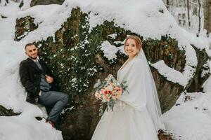 Wedding in winter. Stylish bride in white mink coat and groom in black coat in forest with white snow in cold weather. The bride and groom are walking among rocks and large stones in the forest photo