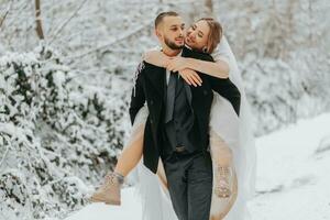 Beautiful wedding couple walking in winter snowy forest, woman in white dress and mink fur coat, bearded man in black coat. The groom carries the bride in his arms, a funny photo