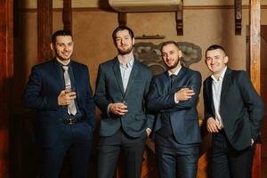 the groom and his friends in stylish suits drink whiskey in the hotel room, the morning before the wedding preparations. Portrait of men drinking whiskey photo