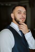 elegante novio con un barba en un hermosa azul chaqueta y blanco camisa. empresario en clásico ropa. un morena hombre prepara para el Boda en el Mañana en su Boda día. foto