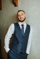 elegante novio con un barba en un hermosa azul chaqueta y blanco camisa. empresario en clásico ropa. un morena hombre prepara para el Boda en el Mañana en su Boda día. foto