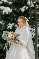 the bride on the background of snowy Christmas trees. Portrait of a beautiful bride in a white poncho and wedding dress in a winter park photo