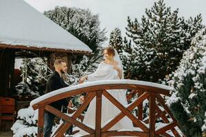 Beautiful and happy bride and groom are walking among snow-covered trees. Groom and bride in the winter park. Bride with a bouquet of flowers in a wedding dress and poncho. Groom in a black coat. photo