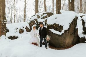 Wedding in winter. Stylish bride in white mink coat and groom in black coat in forest with white snow in cold weather. The bride and groom are walking among rocks and large stones in the forest photo