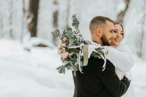 Wedding in winter. The bride and groom are standing against a snowy forest with a blurred background. The bride hugs the groom while holding a bouquet of flowers photo