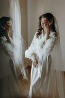 hermosa joven novia con largo Rizado pelo en un de moda blanco túnica en el Mañana. Mañana vendaje de el novia. hermosa joven niña en el Mañana en un blanco vestido. preparación para el Boda foto