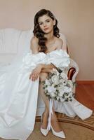 the bride in a wedding dress and a bouquet of flowers in her hands is sitting on the couch. Beautiful model girl with curly hair in a fashionable off-the-shoulder wedding dress photo