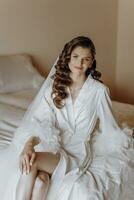 Beautiful young bride with long curly hair in a fashionable white robe in the morning. Morning dressing of the bride. Beautiful young girl in the morning in a white dress. Preparation for the wedding photo