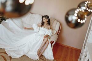 the bride in a wedding dress is sitting on the sofa. Beautiful model girl with long curly hair in a fashionable off-the-shoulder wedding dress photo