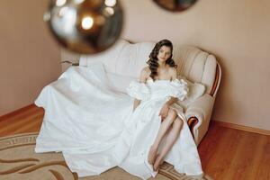 the bride in a wedding dress is sitting on the sofa. Beautiful model girl with long curly hair in a fashionable off-the-shoulder wedding dress photo