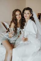 Happy bride and bridesmaid are preparing for the wedding, having fun in pajamas with glasses of champagne in their hands. Support, love and young woman in morning clothes with friend or sister photo