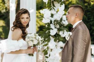 un elegante novia y novio actitud juntos al aire libre en un soleado Boda día en contra un antecedentes de flores en un hermosa ubicación. el novio suavemente abrazos el novia y Besos su. foto