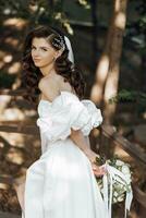 Happy young woman with long curly hair in a white wedding dress, holding a bouquet of flowers, outdoors. Beautiful girl with flowers. Vertical photo