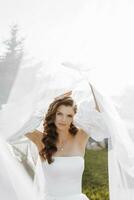 Beautiful bride with long curly hair in a fashionable white wedding dress posing under a veil photo