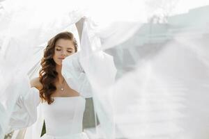 hermosa novia con largo Rizado pelo en un de moda blanco Boda vestir posando debajo un velo foto