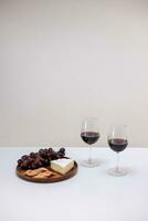 dos lentes de rojo vino, queso y uvas en un blanco mesa foto