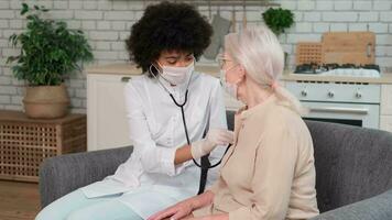 Afro american woman doctor in mask listens to the breathing of an elderly woman at home and supports her holding her hand. Family Doctor, Patient Support, Help at Home, Caring for the Sick. video