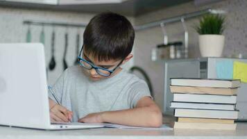 Porträt. das Kind im Brille tut Hausaufgaben mit ein Laptop während Sitzung beim Zuhause im das Küche beim das Tabelle und schreibt mit ein Brunnen Stift im ein Notizbuch. online Technologie video