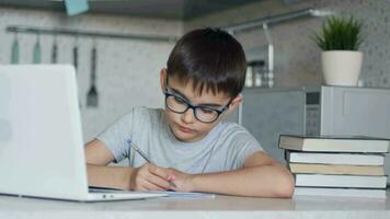 attrayant enfant dans des lunettes enseigne cours tandis que à Accueil séance dans le cuisine à le table en utilisant une ordinateur portable, manuels scolaires, carnet. caméra mouvement vers l'avant video