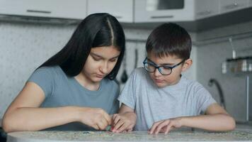 vrolijk mooi jongen en meisje zullen zetten samen een puzzel terwijl zittend Bij een tafel Bij huis in de keuken. detailopname video