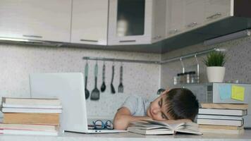 Tired schoolboy fell asleep doing homework sitting at a desk next to a laptop and textbooks. Move the camera to the right. Distance learning video