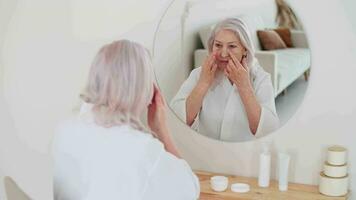 pelle cura, anziano donna, casa cosmetici, anziano età, cosmetico procedure. anziano donna usi viso crema mentre seduta nel davanti di un' specchio video