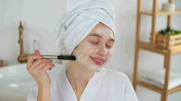 peau se soucier, après bain, produits de beauté à maison, caucasien femme, anti vieillissement. caucasien femme dans une peignoir et une serviette sur sa tête nans une visage masque avec une brosse video