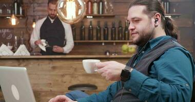 Confident man engaged in a video call enjoys a coffee in a small restaurant. Freelancer with long hair having a video conference.