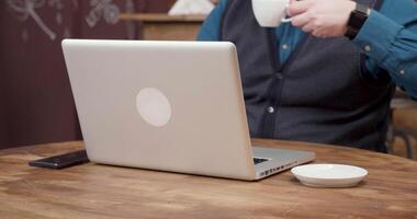 Male freelancer sips coffee while working from a small coffee shop. Close up view of a laptop, coffee and smartphone on wooden table. video