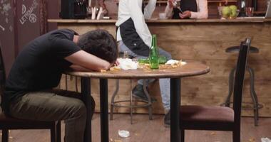 Waiter in black apron tries to wake a drunk customer that fell asleep on a dirty table. closed restaurant after a busy day. video