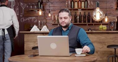 bonito homem com grandes cabelo olhando às dele computador portátil tela e bebidas café. jovem homem de negocios assistindo uma vídeo em dele computador em uma café pausa. video