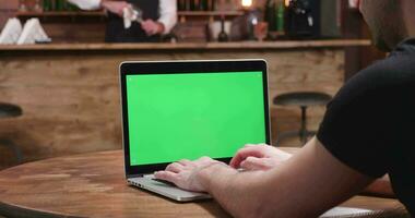 ung man i tillfällig kläder typer snabb på en bärbar dator med grön skärm. han är i en årgång pub på de tabell. en bartender är arbetssätt i de bakgrund. video