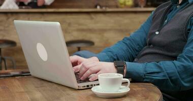 detalhe tiro do masculino mãos digitando uma texto em dele computador portátil e bebidas café. homem trabalhando em dele computador a partir de uma restaurante. video