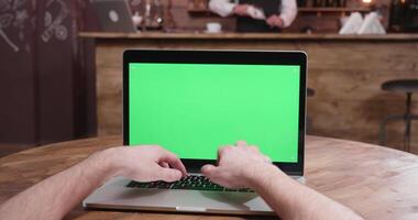 pov parallasse tiro di uomo mani digitando su un' computer con verde schermo nel un' accogliente bar mentre il barista è Lavorando nel il sfondo a il bar contatore. video