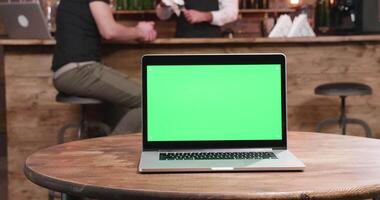 Modern laptop with green screen on a table in a vintage restaurant. A customer and bartender are in the background chatting video