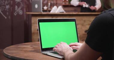 uomo digitando veloce su un' il computer portatile con verde schermo. lui si siede a il tavolo nel un' Vintage ▾ fricchettone pub mentre il barista è Lavorando a il bancone da bar video