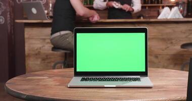 Zoom in shot on laptop with green screen on a table in a vintage cafe, pub or restaurant. In the background a customer is chatting with the bartender video