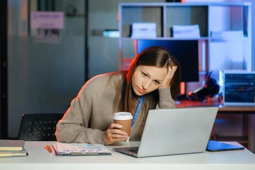 negocio mujer Bebiendo café a obtener algunos energía para trabajando tiempo extraordinario sentado a escritorio utilizando computadora y haciendo tiempo extraordinario foto