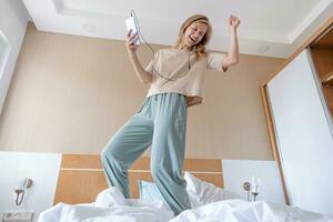 mujer saltando en cómodo acogedor cama a hotel habitación escuchando a el música, disfrutando Mañana foto