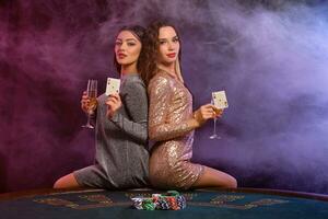 Two girls are playing poker at casino, holding glasses of champagne and cards, sitting on table with chips on it. Black, smoke background. Close-up. photo